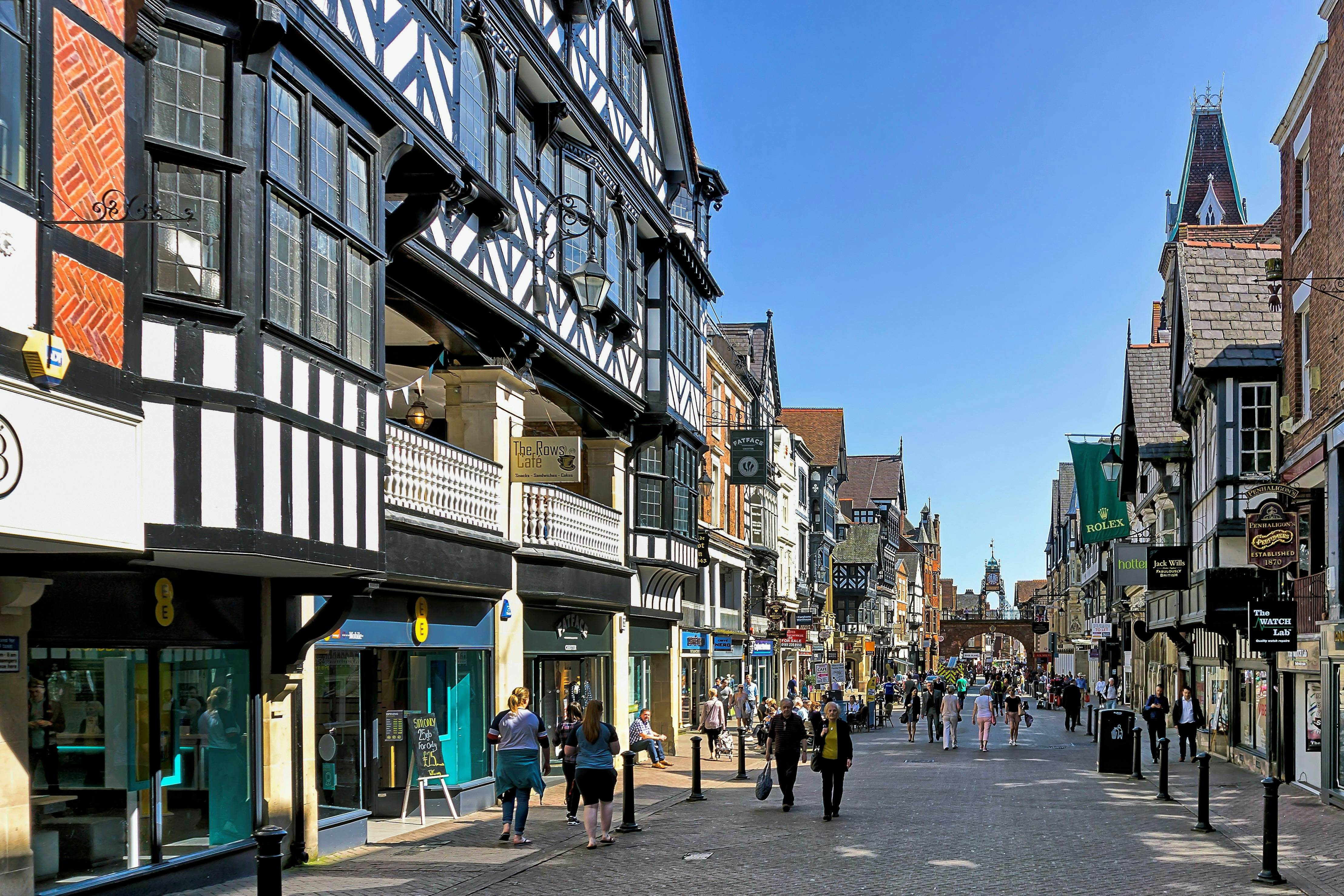 View along the main street in the centre of Chester, Cheshire, UK.  Shops can be seen on either side of the road and people can be seen walking and sitting on benches. The rows of upper level shops are pictured on left. 
